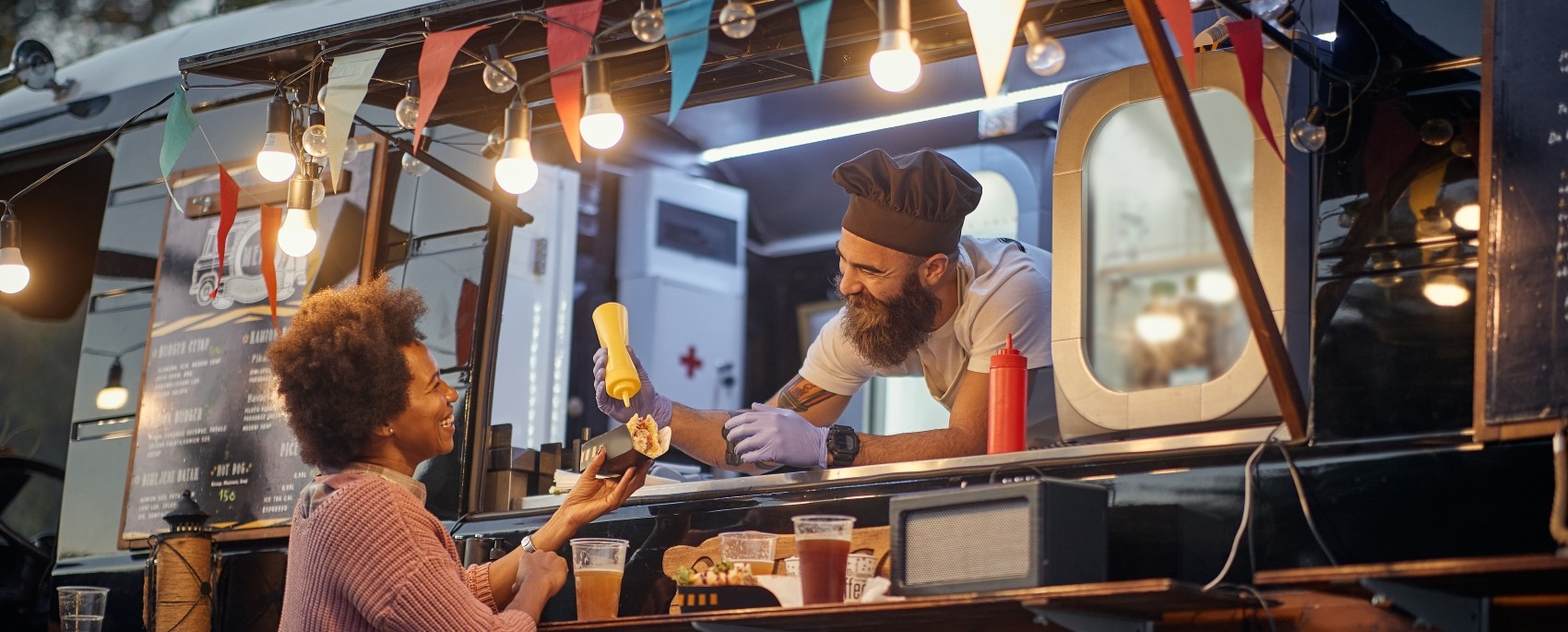 A man operating a mobile food truck assists a woman by helping her put mustard on her hot dog