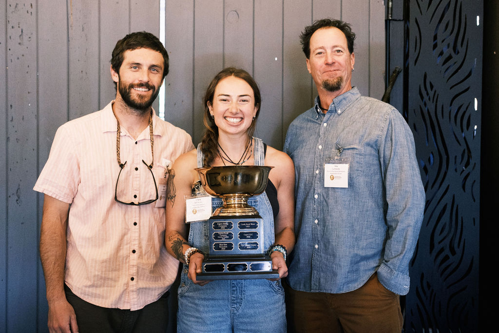 A woman and two men pose for a photo together. The woman is standing between the two men, holding a large trophy