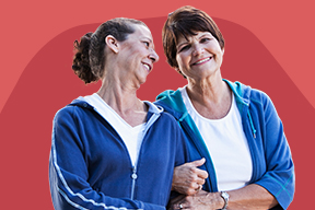 Two older women link arms - one smiles at the other and the other smiles at the camera