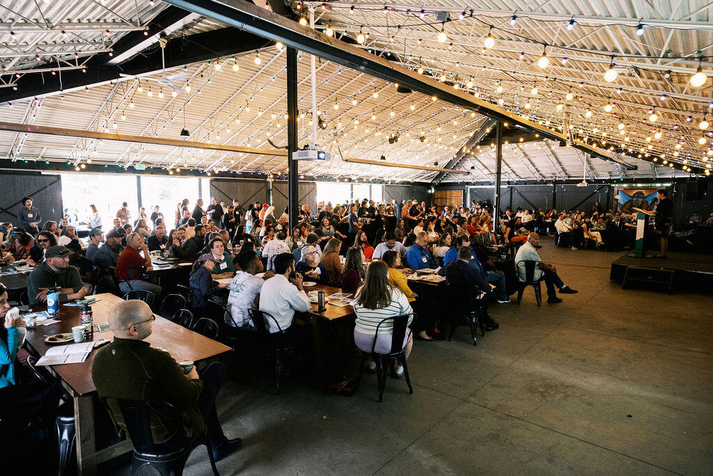 People watch a presentation, while sitting at tables, inside a large tent-like structure