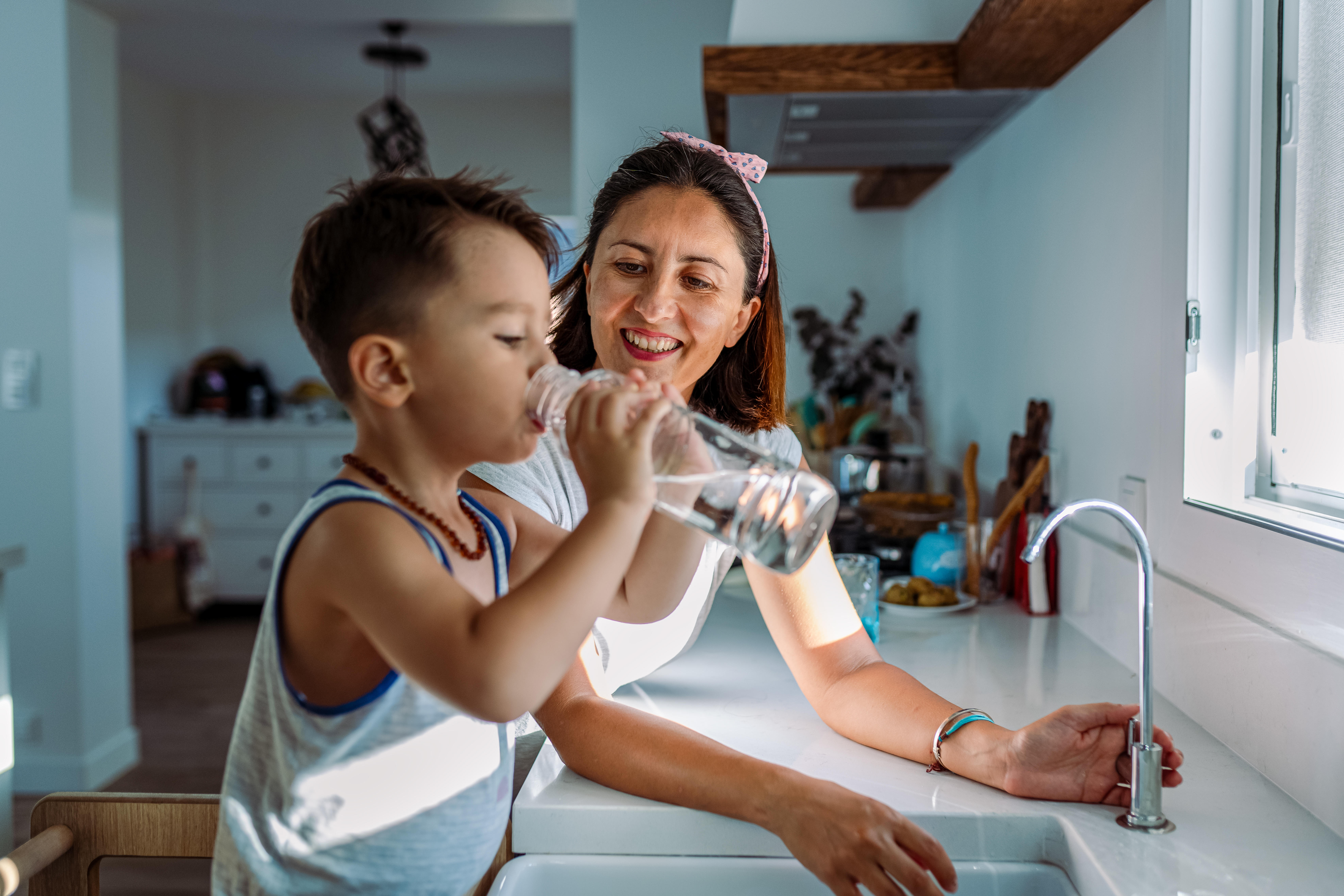 Latino mom and young boy at the kitchen sink drinking a glass of water. 