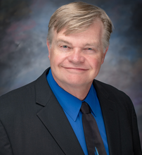 Portrait image of Scott Linder in a dark blue with a light blue shirt and dark tie smiling.