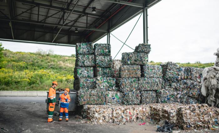 Male and female waste management workers in protective suits and hardhats standing outdoors next to stacks of compacted and bundled recyclables.