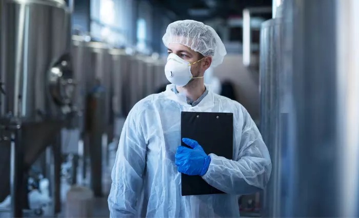Technologist in protective white suit with hairnet and mask standing in food factory.
