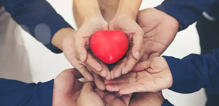 multiple peoples' hands stacked on each other supportively holding a heart