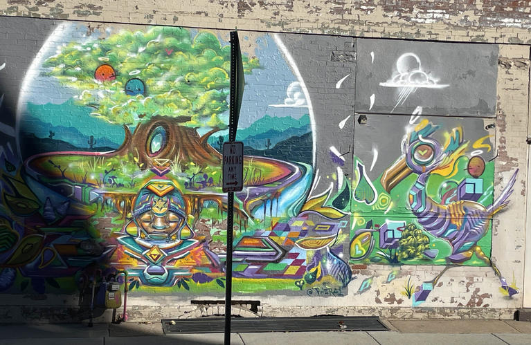 Colorful abstract mural on a public brick wall including mountains, tree, angels, water, and other animals as well as a single person.