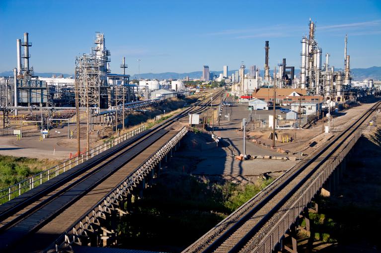 Photo of train tracks passing through an industrial oil refinery with Denver skyline in the background. (Suncor oil refinery)