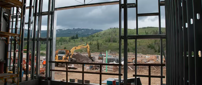 View of construction site with large construction equipment from inside the frame of a building with cloudy skies and mountains in background.