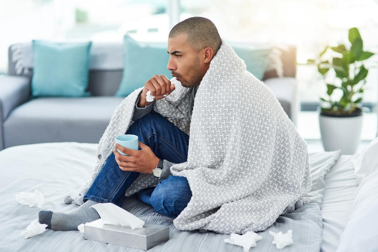 man sitting on bed wrapped in blanked coughing
