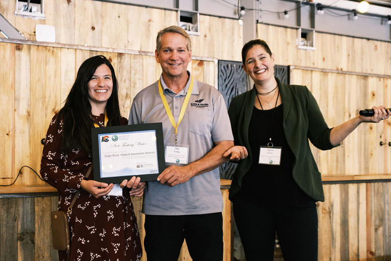 Two representatives from Eagle River Water & Sanitation District stand with CGBN Manager, Rayna Oliker, with their framed awarded certificate for the winning the Best Tasting Water.
